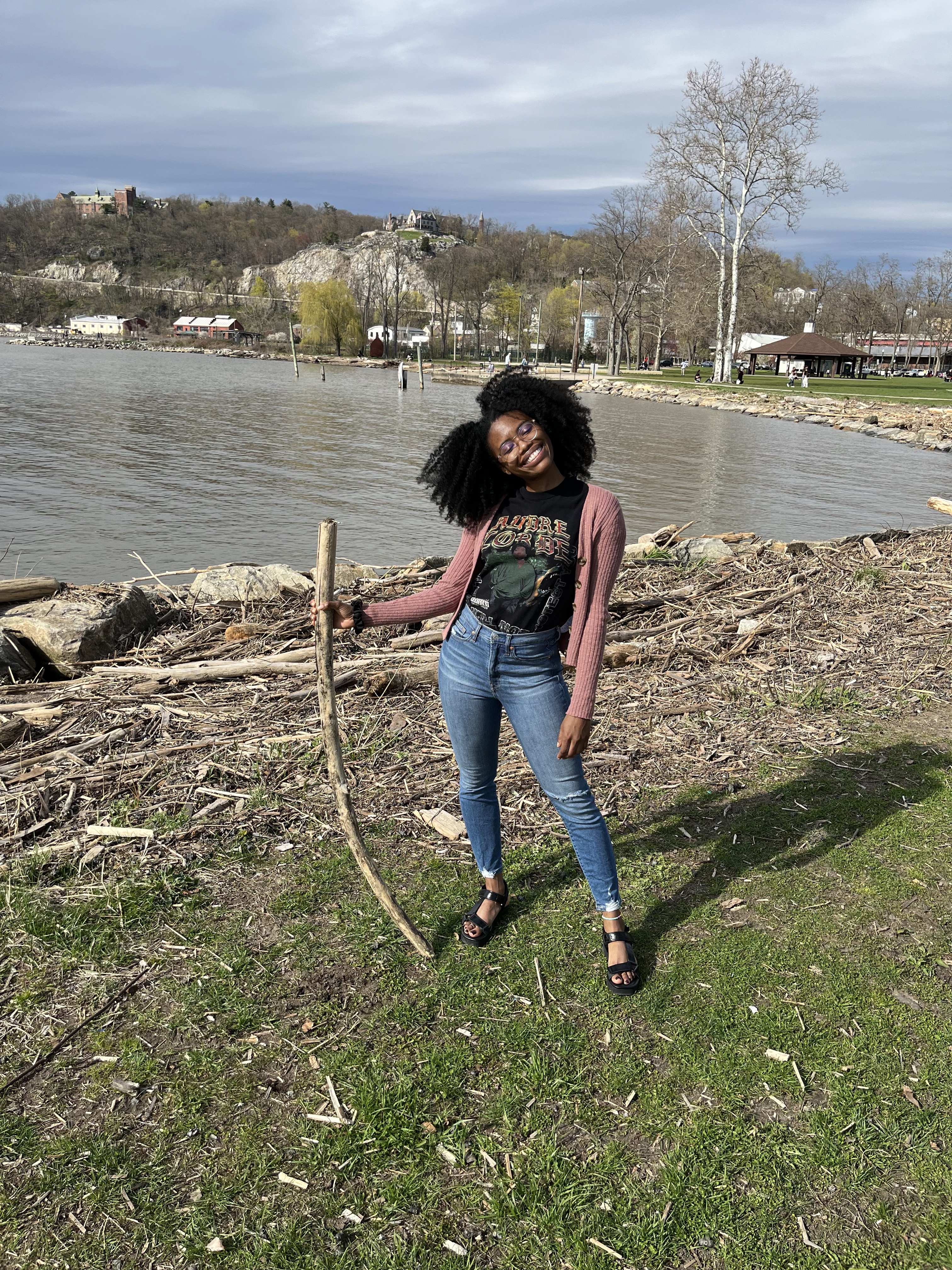 A picture of Dominique, a Black woman wearing jeans and a pink cardigan sweater and a Toni Morrison t-shirt, standing in front of a pond holding a stick.